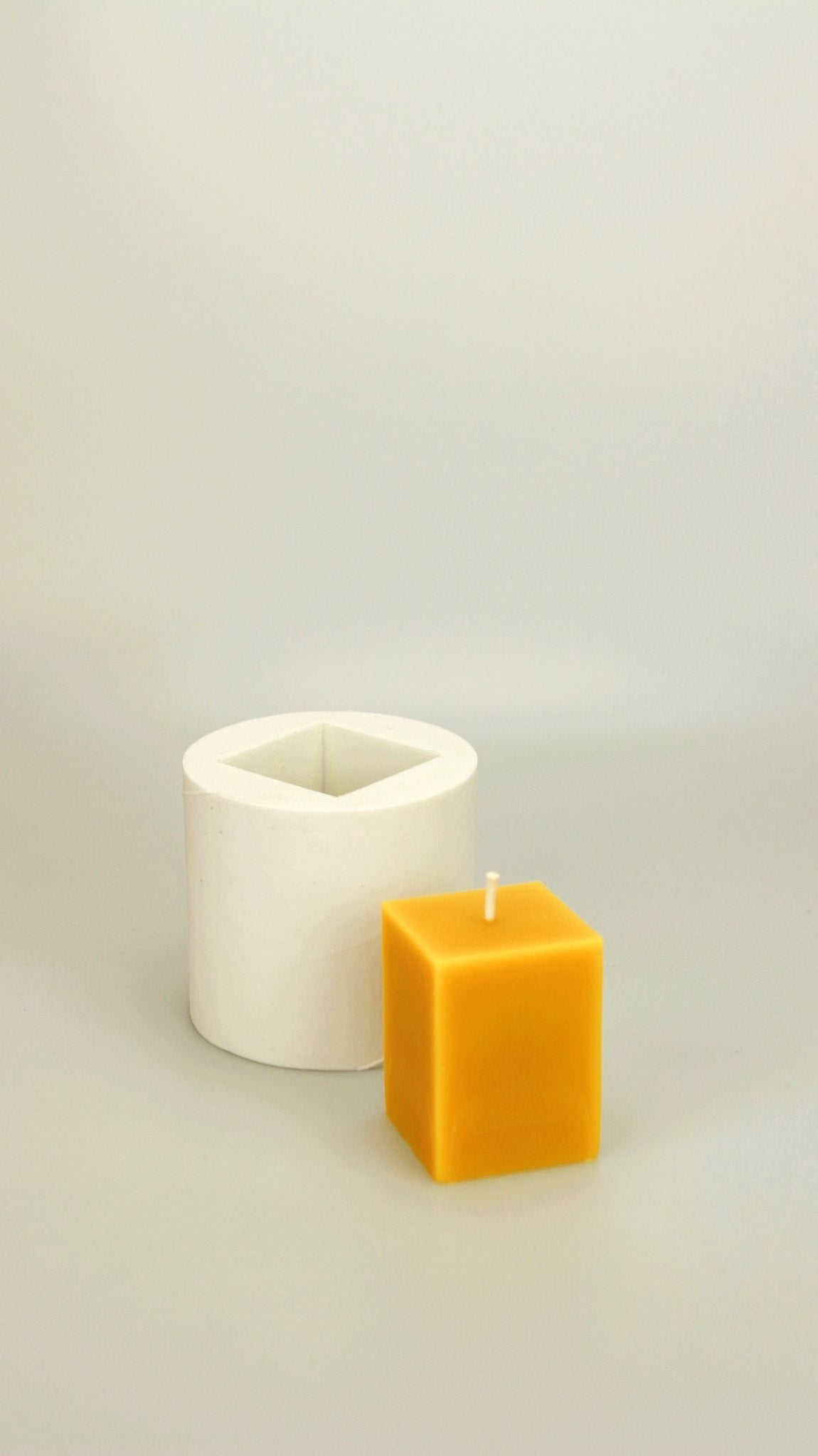 Small Cubes beeswax candle next to its short white silicone mould with a square cavity, cream background